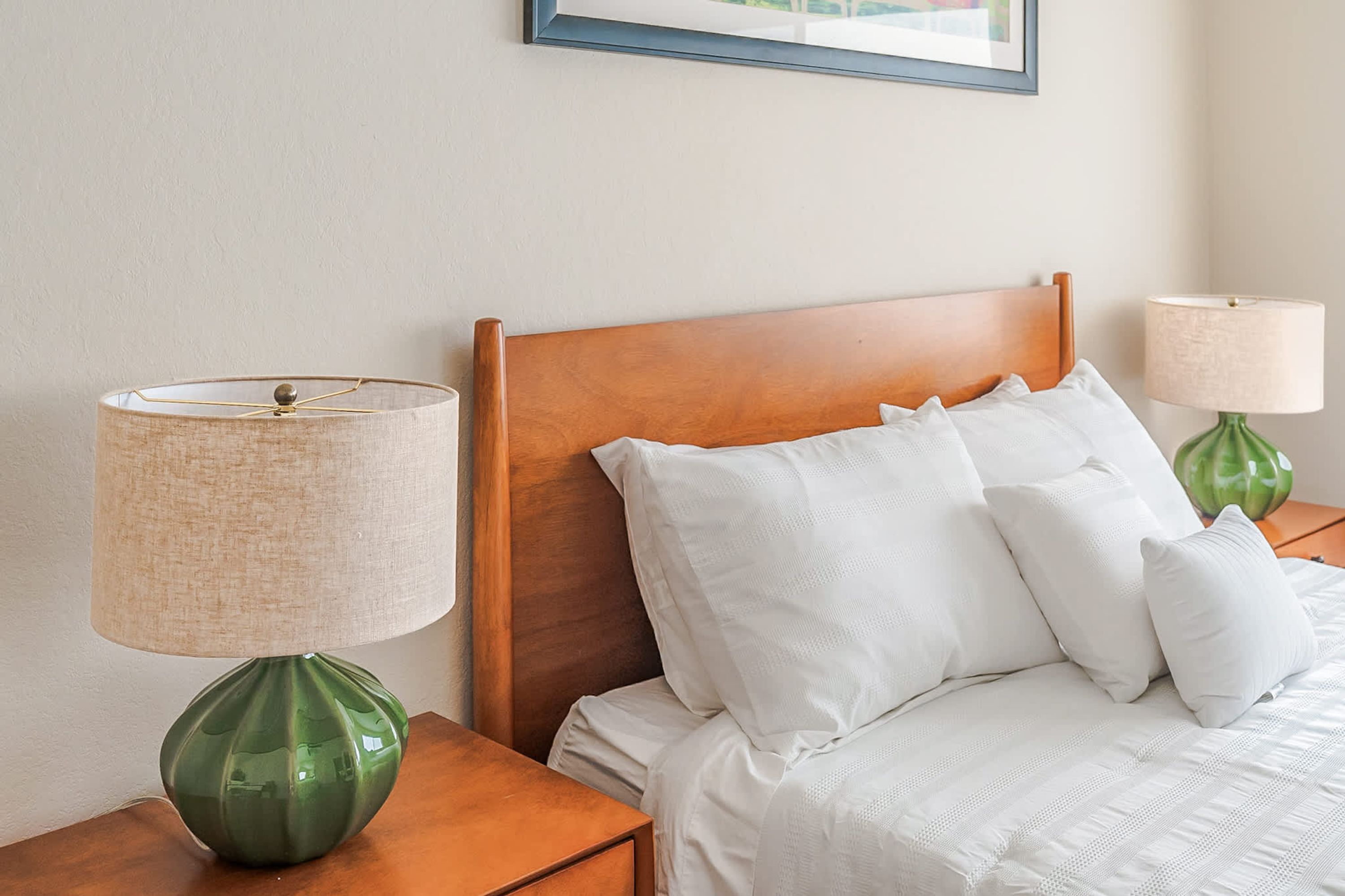Una cama bien tendida con sábanas y almohadas blancas, con dos lámparas verdes a cada lado sobre mesitas de noche de madera.