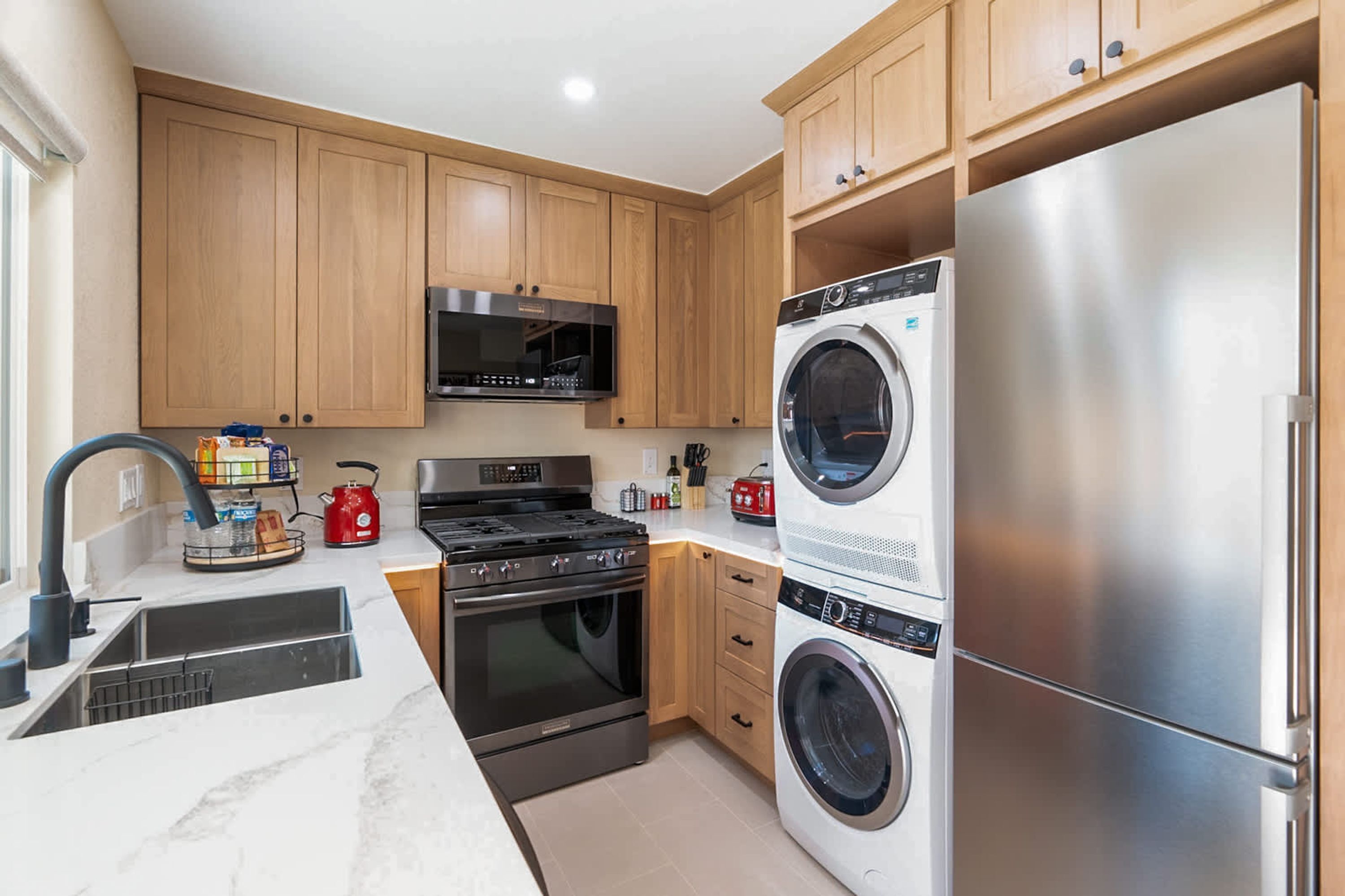 A modern kitchen with wooden cabinets, a stainless steel refrigerator, a built-in oven, a microwave, and a washing machine.