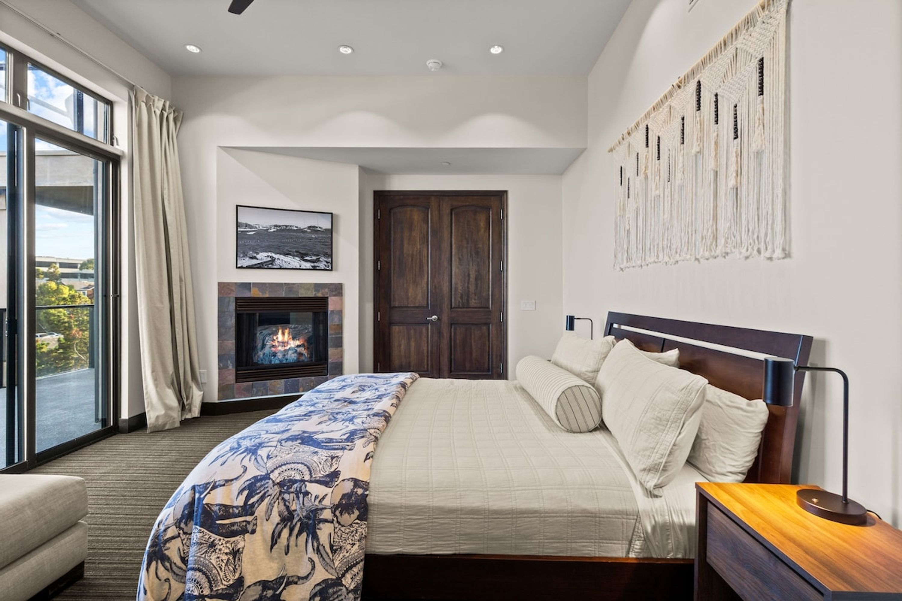 A modern bedroom with a large bed, a fireplace, a view through a sliding door, and a macrame wall hanging.