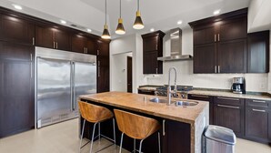 A modern kitchen with dark wood cabinets, stainless steel appliances, a central island with stools, and pendant lighting. - Dana Point Luxe Str14-007 (Dana Point)