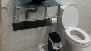 Un baño con pared de azulejos blancos, un lavabo negro con un espejo encima, un inodoro y un cubo de basura.