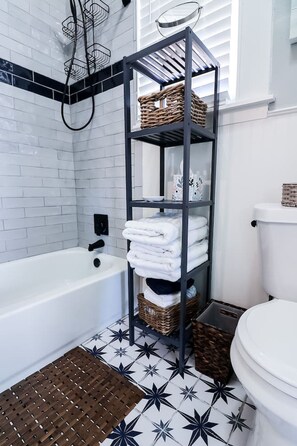 A bathroom with a white bathtub, a black metal shelf with baskets, and a toilet. - Vibrant Bungalow 2 Minutes to the Beach (Galveston)