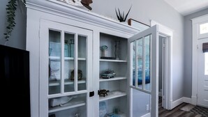 A white built-in cabinet with multiple glass doors, shelves with various items, and a partially open door revealing a hallway. - Vibrant Bungalow 2 Minutes to the Beach (Galveston)