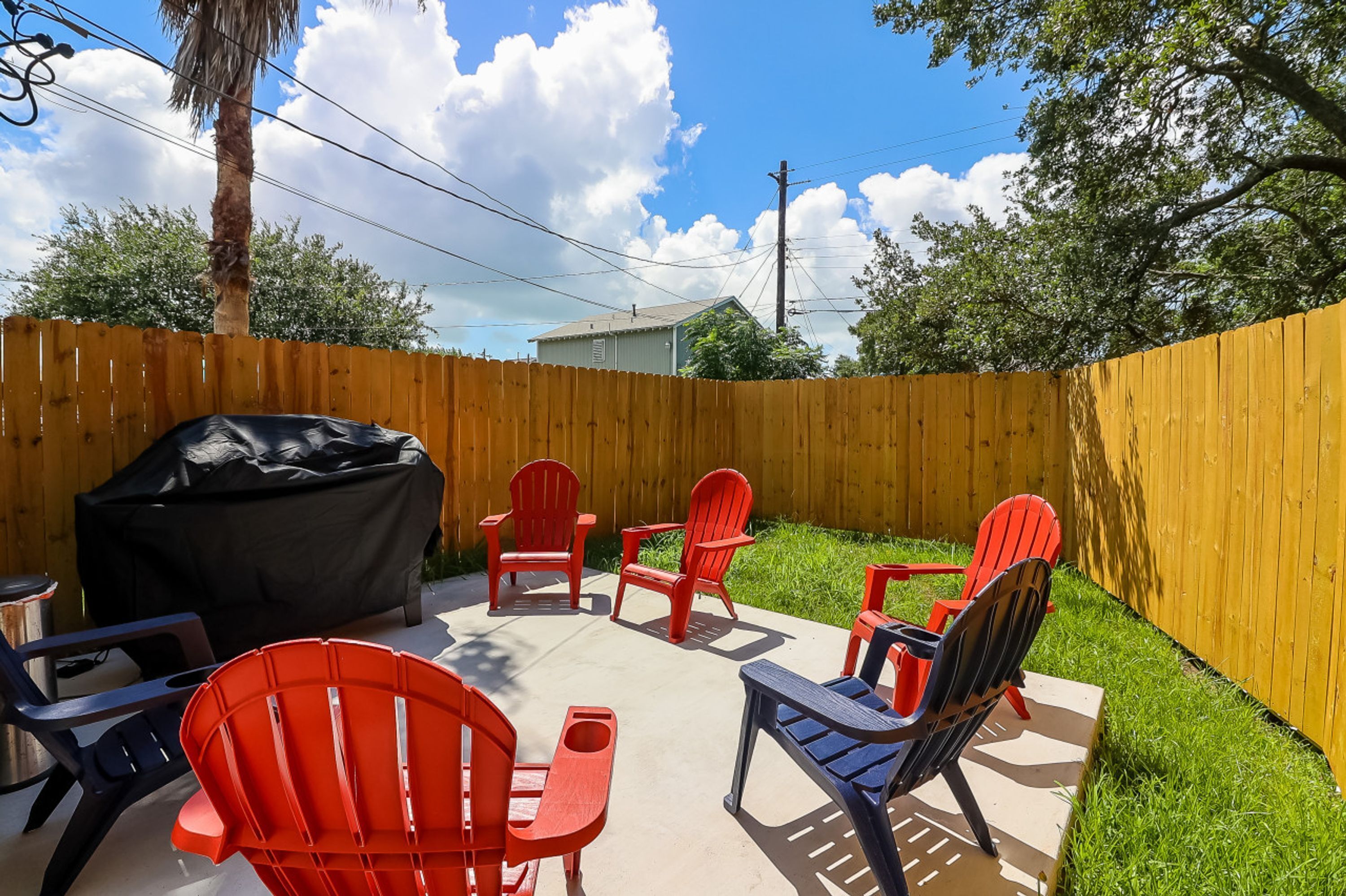 A backyard with a wooden fence, a covered grill, and red Adirondack chairs arranged around a concrete patio.
