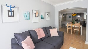 Ein modernes Wohnzimmer mit einem dunkelgrauen Sofa, pinken Kissen und gerahmten Kunstwerken an der Wand. Im Hintergrund ist ein Essbereich mit einem Holztisch und Stühlen zu sehen.