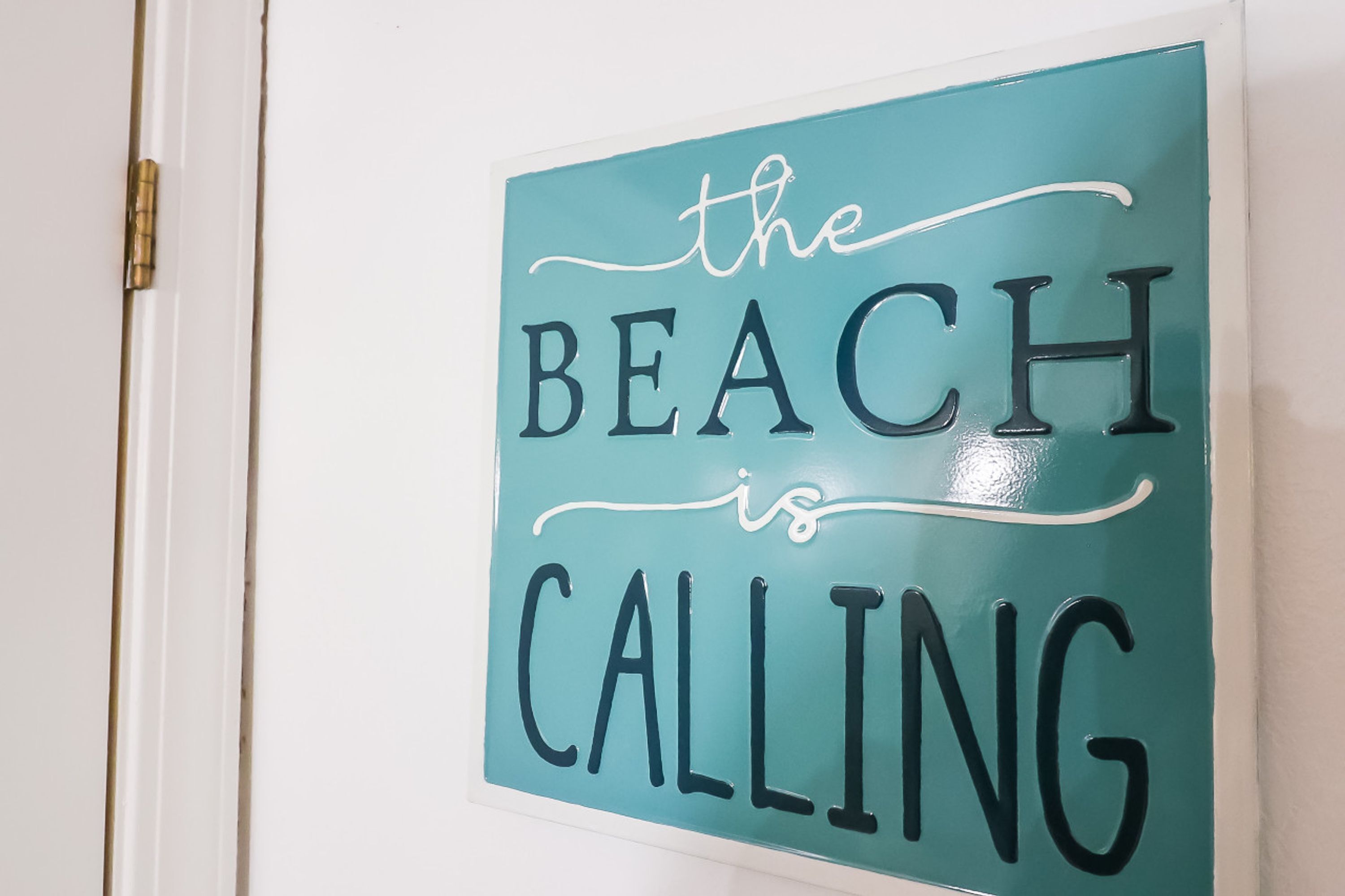 Un letrero azul turquesa con la inscripción 'the BEACH is CALLING' en una puerta blanca.