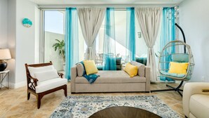 A living room with a sofa, a wooden chair, a hanging chair, a clock, and a view of the outdoors. - Warm and Welcoming Views at Diamond Beach (Galveston)