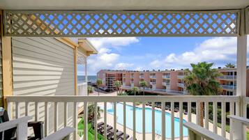 A balcony with a view of a swimming pool, palm trees, and a hotel building in the background.