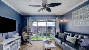 A living room with a black leather sofa, a white coffee table, a ceiling fan, a TV mounted on the wall, and a balcony with a view of buildings. - Beautiful Ocean Views at The Victorian (Galveston)