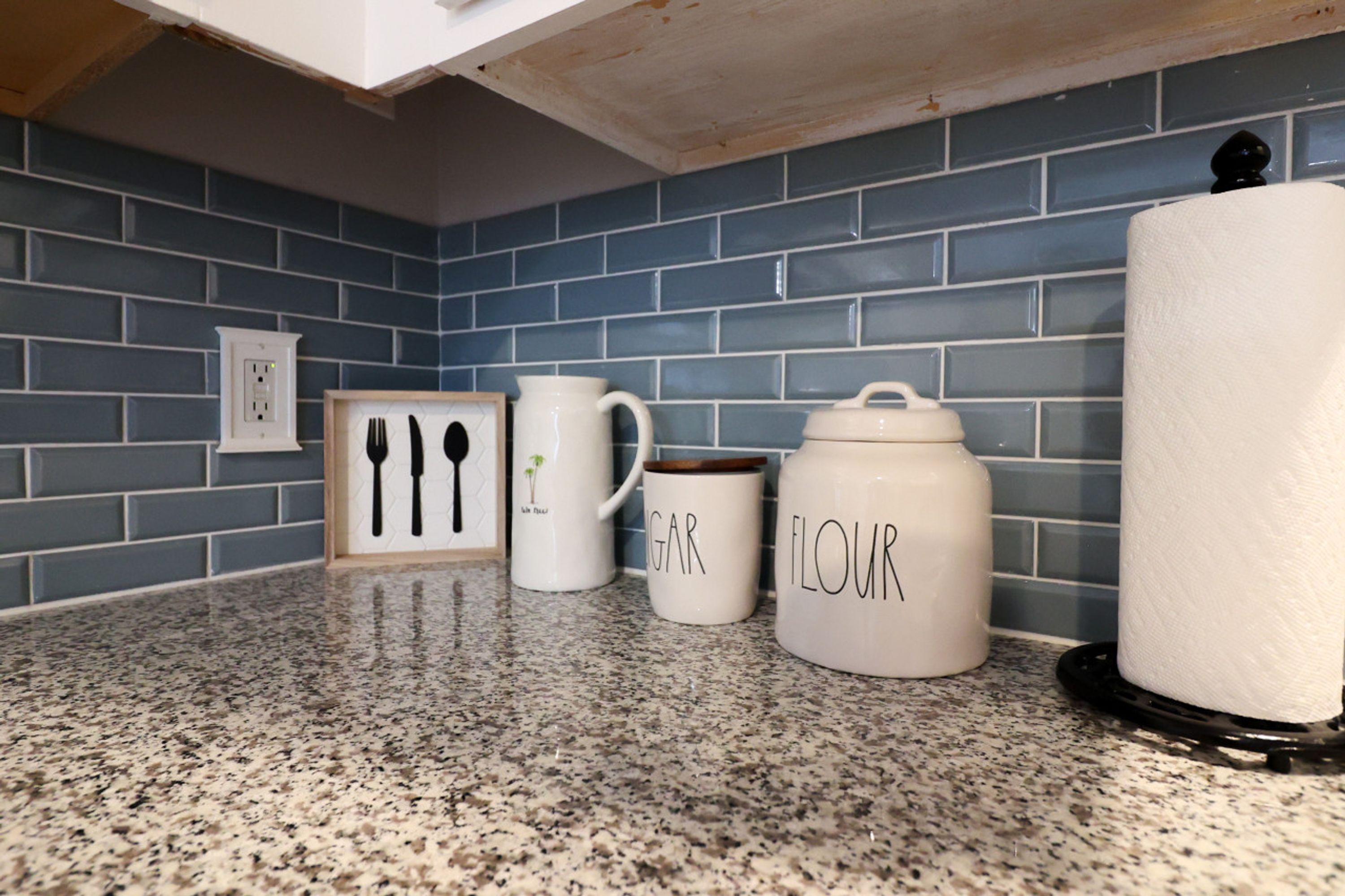 A kitchen counter with three white ceramic containers labeled "GIRL FRIENDS," "GAR," and "FLOUR," and a white paper towel holder.