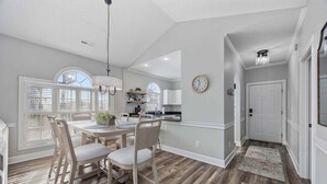 A dining area with a wooden table and chairs, a kitchen in the background, and a clock on the wall. - Wild Iris Condo - Cozy Myrtle Beach Retreat (Myrtle Beach)