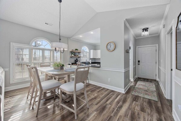 A dining area with a wooden table and chairs, a kitchen in the background, and a clock on the wall.