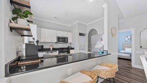 A modern kitchen with white cabinets, a black countertop, and a built-in microwave. There are two wicker bar stools and a potted plant on the counter. - Wild Iris Condo - Cozy Myrtle Beach Retreat (Myrtle Beach)