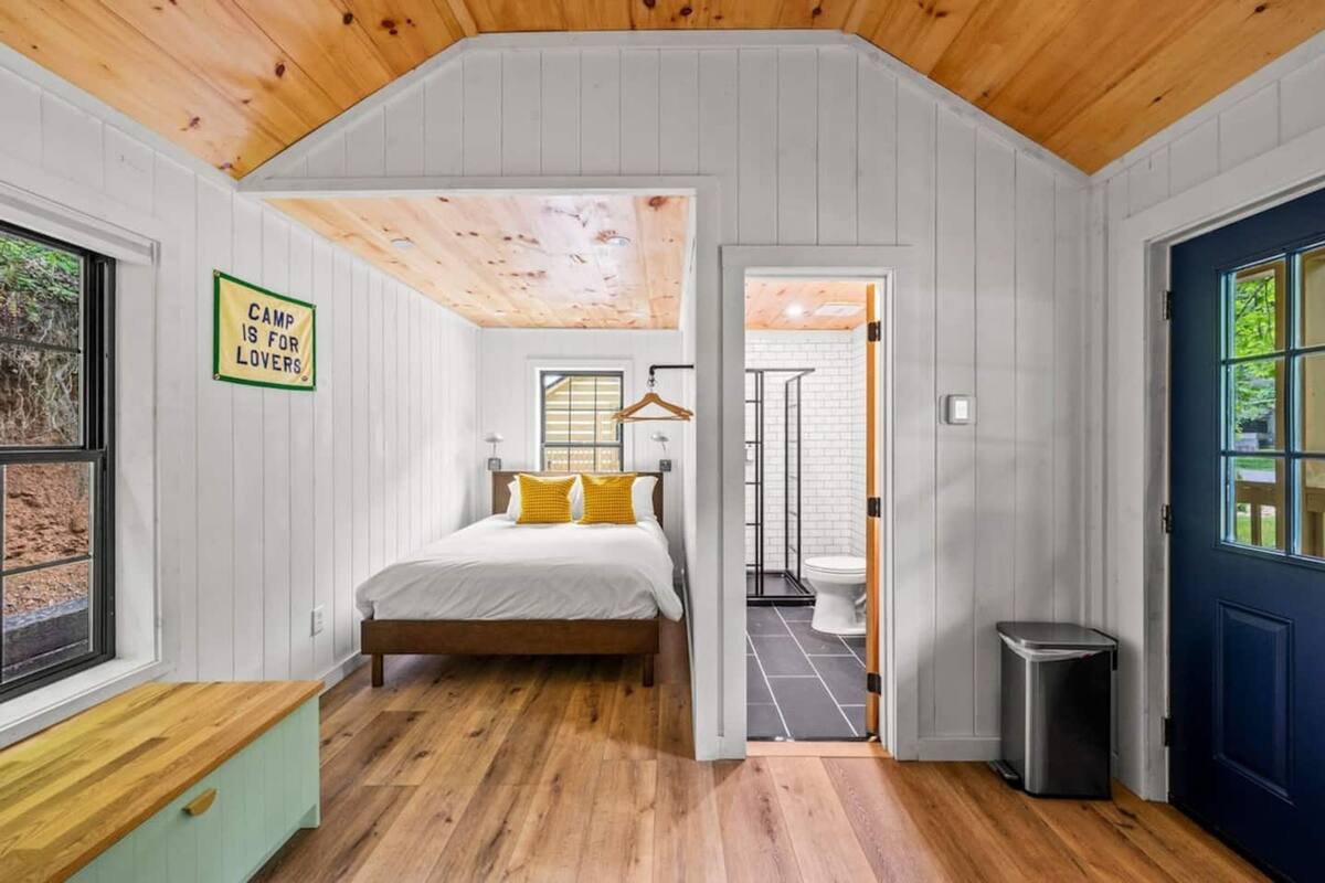 A bedroom with a bed, a wooden ceiling, a window, and a door leading to a bathroom.