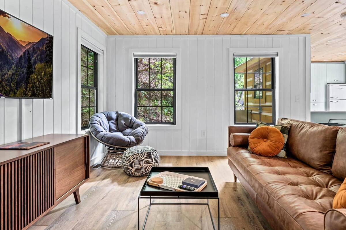 A modern living room with a wooden ceiling, a large TV, a leather sofa, a brown couch, and a wooden cabinet.