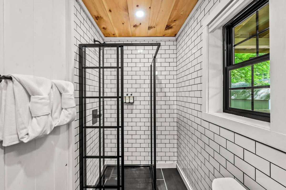 A bathroom with white tiled walls, a wooden ceiling, a black metal shower enclosure, and a window with a view of greenery.