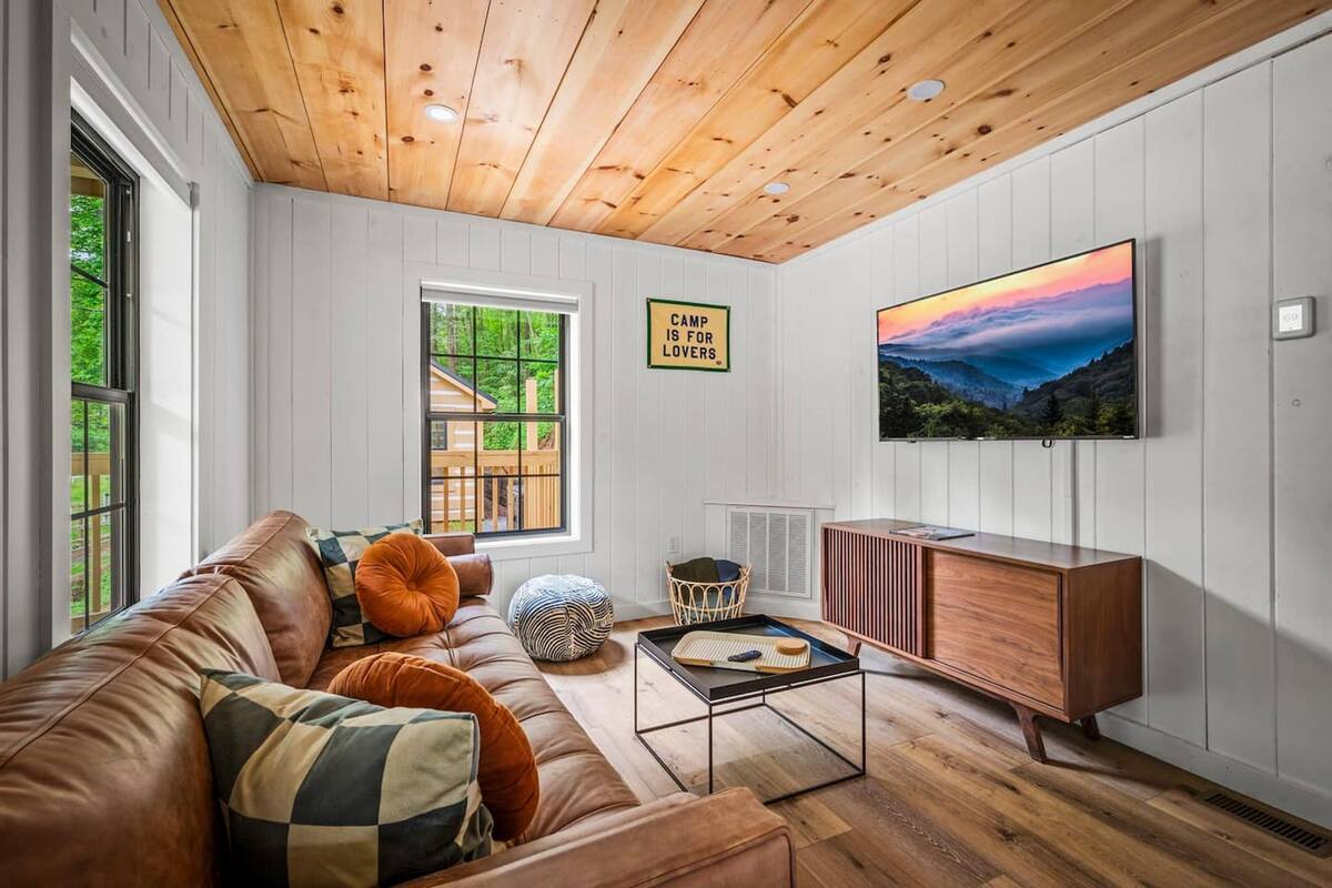 A cozy living room with a wooden ceiling, a large TV, a leather couch, and a view of the outdoors through a window.