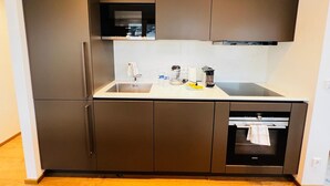 Una cocina compacta con armarios oscuros, un refrigerador de acero inoxidable, un horno empotrado y una pequeña encimera con un lavamanos y varios utensilios de cocina.