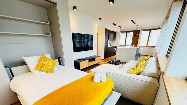 Una habitación de hotel moderna con dos camas, un sofá, un televisor de pantalla plana, una pequeña cocina y ventanas amplias.
