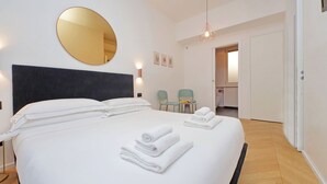 Une chambre d’hôtel avec un grand lit, un miroir rond, une petite table avec des chaises et une porte donnant sur une autre pièce.