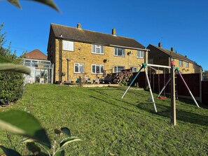 A residential area with a two-story house, a grassy yard, and a swing set. - NEW Cosy 2 Bedroom Home | Free Parking + Garden (Canterbury)