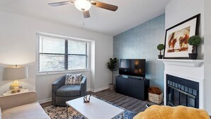 Ein modernes Wohnzimmer mit Deckenventilator, Flachbildfernseher, Kamin und einem großen Fenster mit Jalousien.