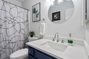 A bathroom with a white toilet, a sink with a mirror above it, a shower curtain with a floral pattern, and a small potted plant on the sink's edge. - Charming Private Oasis - close to airport, mall, and downtown. (Raleigh)