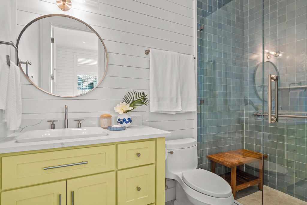 A bathroom with a white sink, green cabinets, a round mirror, a toilet, and a glass shower enclosure.