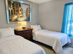 Une chambre d’hôtel avec deux lits, un tableau représentant un paysage de canal, une lampe et une fenêtre avec des rideaux bleus.