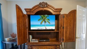A wooden entertainment center with a television displaying a beach scene, a small table with books, and a chair. - 4-bedroom in at the Top Notch Villa in  Deltona, 25 min to Disney and Universal. (Deltona)