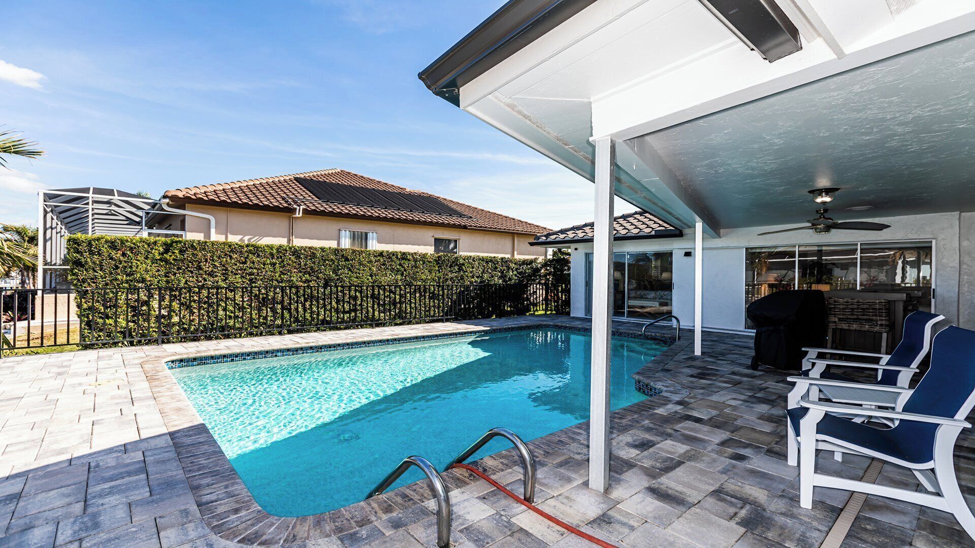 A swimming pool with a tiled deck, a covered patio area with chairs, and a house with a tiled roof in the background.