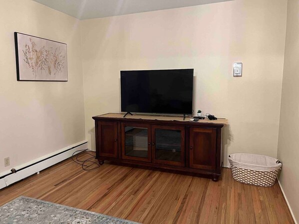 A living room with a flat-screen TV on a wooden cabinet, a framed picture on the wall, and a laundry basket.