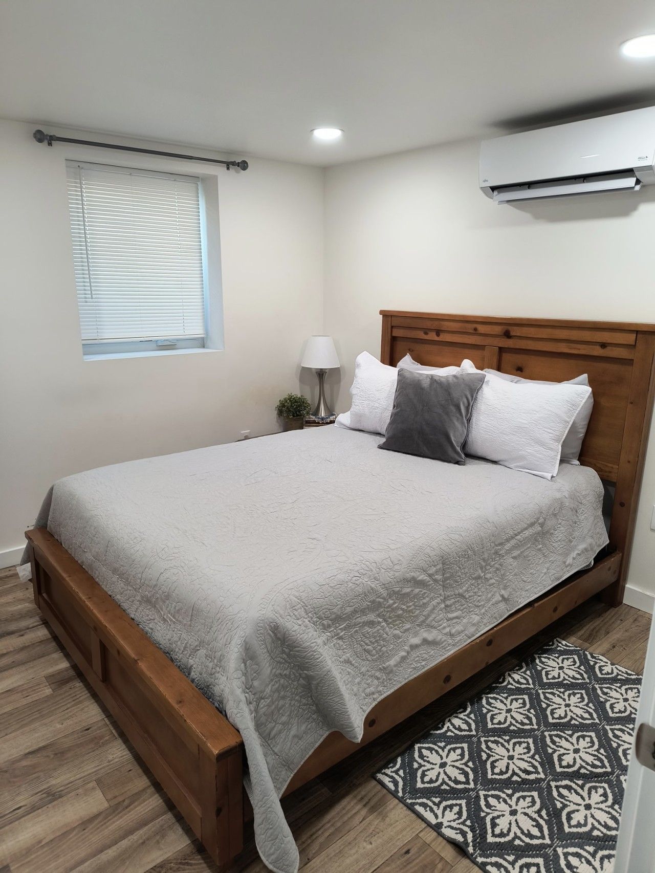 Un dormitorio con una cama de madera, una ventana con persianas, una lámpara de noche y un equipo de aire acondicionado.