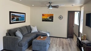 Ein Wohnzimmer mit einem grauen Ecksofa, einem kleinen Hocker, einem Deckenventilator und zwei gerahmten Bildern an der Wand.