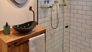 Un baño con tocador de madera, un espejo redondo, un lavamanos tipo cuenco negro y una ducha con mampara de vidrio y ventana.