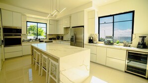 Une cuisine moderne dotée d’un grand îlot central, d’appareils électroménagers en acier inoxydable et offrant une vue sur l’océan grâce à de grandes fenêtres.