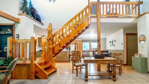 A spacious living area with a wooden staircase, a dining table with chairs, and a kitchen in the background.