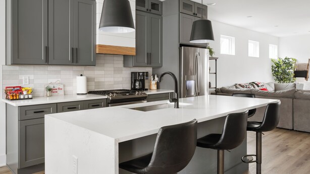 A modern kitchen with grey cabinets, a white countertop, and a stainless steel refrigerator. There are two black bar stools and a large island with a sink. - The Linden Home - Fountain Square Indianapolis (Indianapolis)