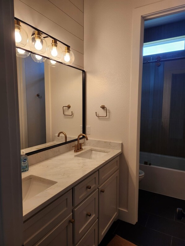A bathroom with a white vanity, a large mirror, a sink with a gold faucet, and a shower area visible through a glass door. - Hillside Hideaway (Bokchito)