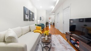 A modern living room with a sofa, a coffee table, a TV, and a bookshelf. - Private Patio Apt • Min to NYC • Sleeps up to 9 • Free Parking • Washer/Dryer (Hoboken)