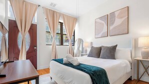 A well-lit bedroom with a large bed, a bedside table, a lamp, and a view of the outdoors through a window. - Private Patio Apt • Min to NYC • Sleeps up to 9 • Free Parking • Washer/Dryer (Hoboken)