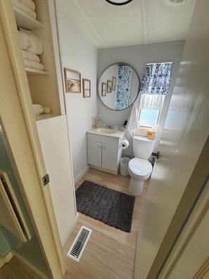 A compact bathroom with a toilet, sink, and a mirror. There is a towel rack with towels, a small cabinet, and a window with a floral curtain.