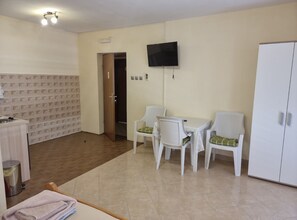 Une pièce avec un sol carrelé, un téléviseur fixé au mur, une armoire blanche et une petite table avec deux chaises.