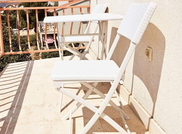 Un balcon avec une chaise pliante blanche et une petite table, une rambarde en métal, et une vue sur un immeuble et des arbres.