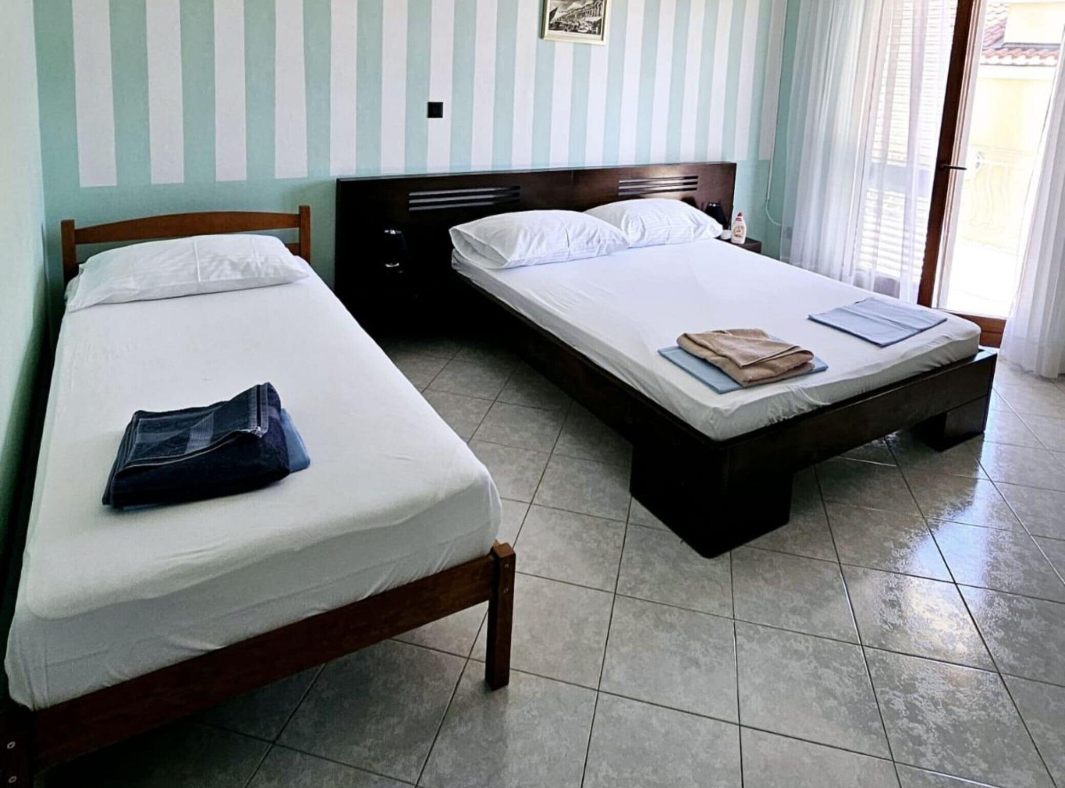 Une chambre avec deux lits, une tête de lit en bois, un sol carrelé et une fenêtre avec des rideaux.