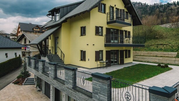 A multi-story yellow building with a balcony, a stone facade, and a paved driveway.