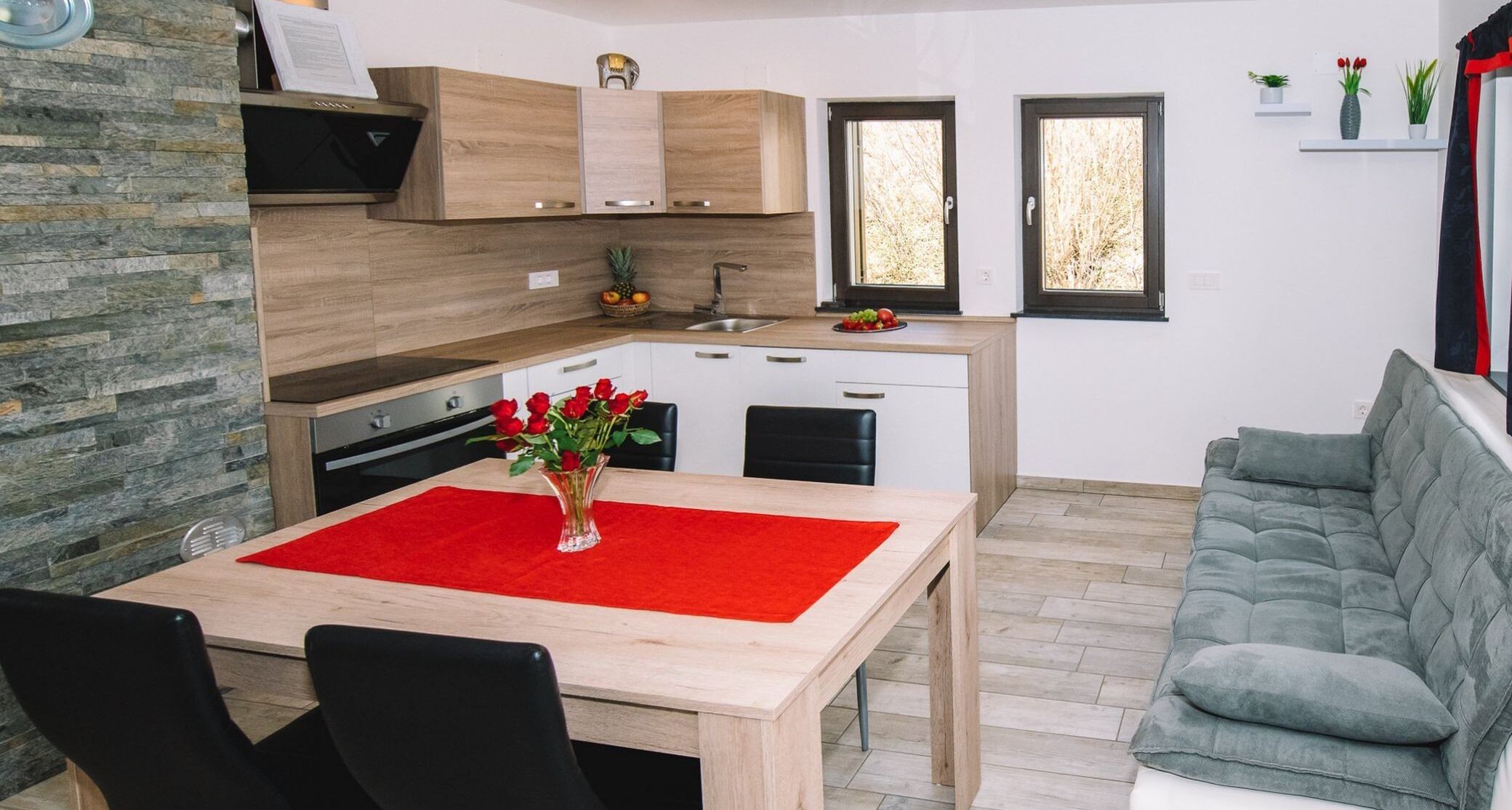 A modern kitchen with wooden cabinets, a dining table with a red placemat, and a grey sofa.