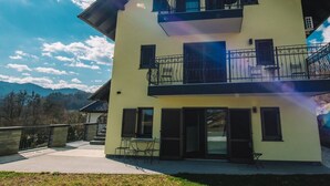 A two-story building with a balcony, a patio area with outdoor seating, and a view of mountains in the background. - Bled Prestigious and Stylish Apartments - Standard One Bedroom Apartment with Balcony (Bled)
