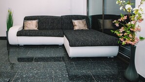 A modern, L-shaped sectional sofa with two beige cushions in a minimalist room with dark tiled flooring. - Bled Prestigious and Stylish Apartments - Standard One Bedroom Apartment with Balcony (Bled)