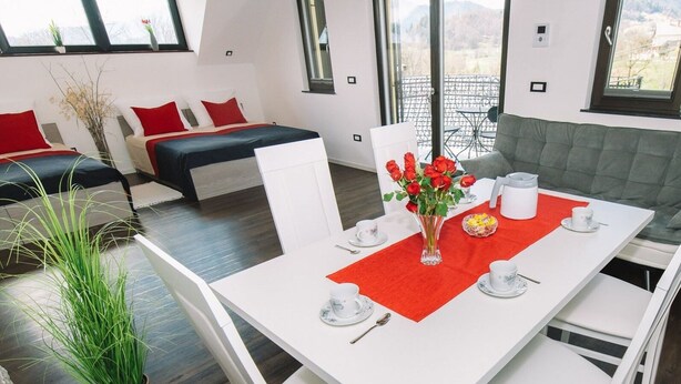 Un coin repas moderne avec une table blanche dressée pour quatre personnes, entourée de chaises blanches. Un chemin de table rouge est posé sur la table, et un vase avec des fleurs rouges se trouve au centre.